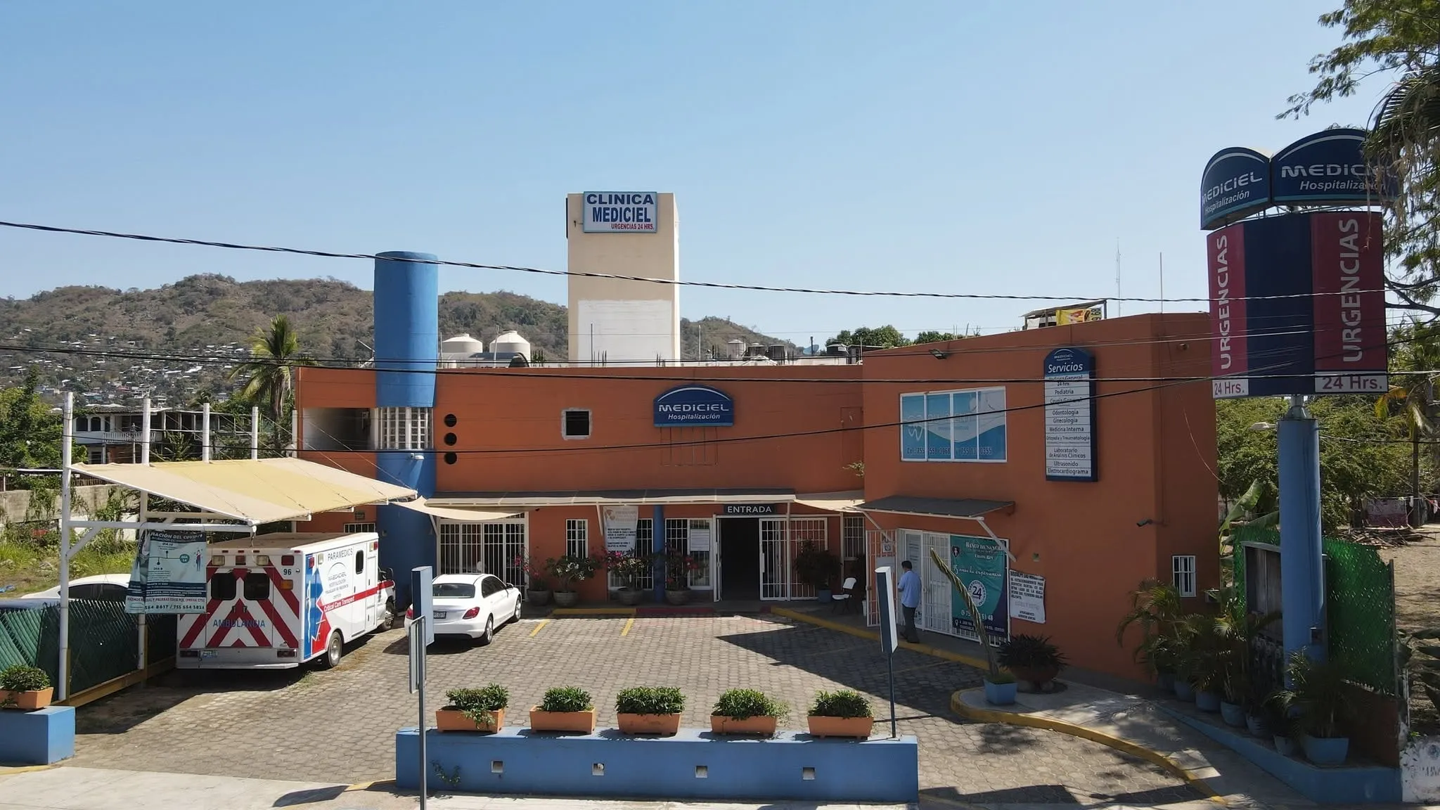 Vista aérea de la fachada de Clínica Mediciel: edificio color terracota con el rótulo de Urgencias 24 horas, y una ambulancia estacionada junto a la entrada.
